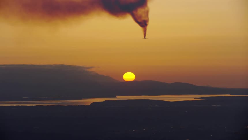 Flying airplane leaves a dark trail of smoke in an orange sky with a bright sun on the horizon performing acrobatic maneuvers