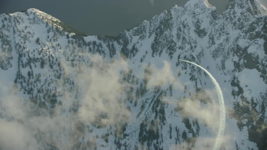 An acrobatic single engine airplane emerges from behind a golden cloud with smoke and does a loop revealing snow covered mountains in this stunning shot