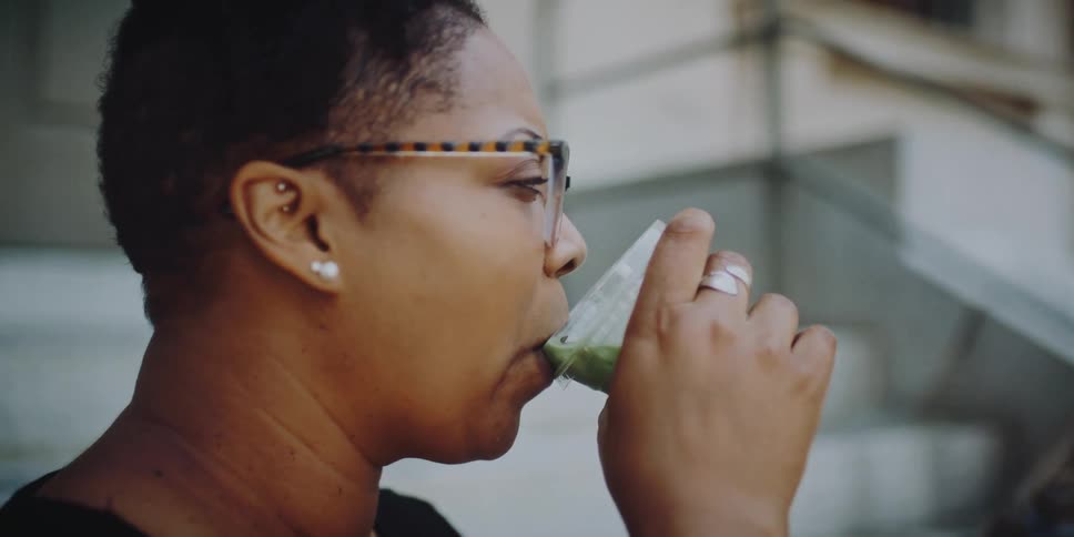 Woman drinking green juice from a plastic cup while talking to a friend on the stairs of a building