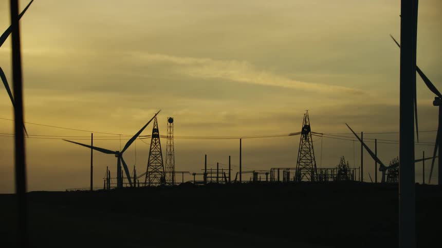 Wind turbines slowly turn blades on wind farm as the camera rotates around a power sub station that distributes the generated electricity