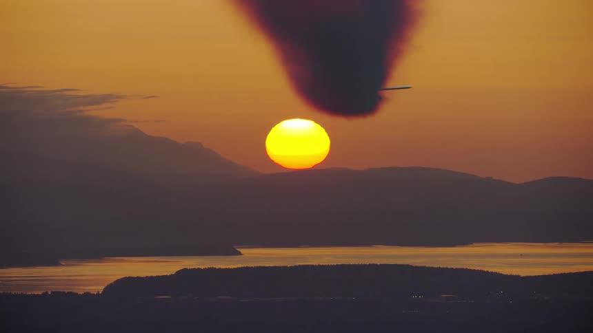 Smoke billows out of the back of a single engine airplane as it flies towards the setting sun