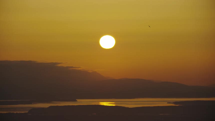 Wide shot of a setting sun over the Pacific Ocean as a small single engine aircraft turns sharply for landing at the airport