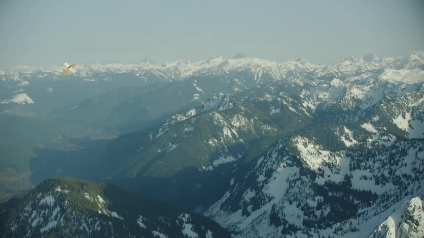 A single engine airplane maneuvers in a wide open sky with huge mountains and deep fjords in the background