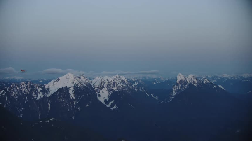Blue Hour shot of a small orange acrobatic airplane flying in front of dramatic snow covered peaks