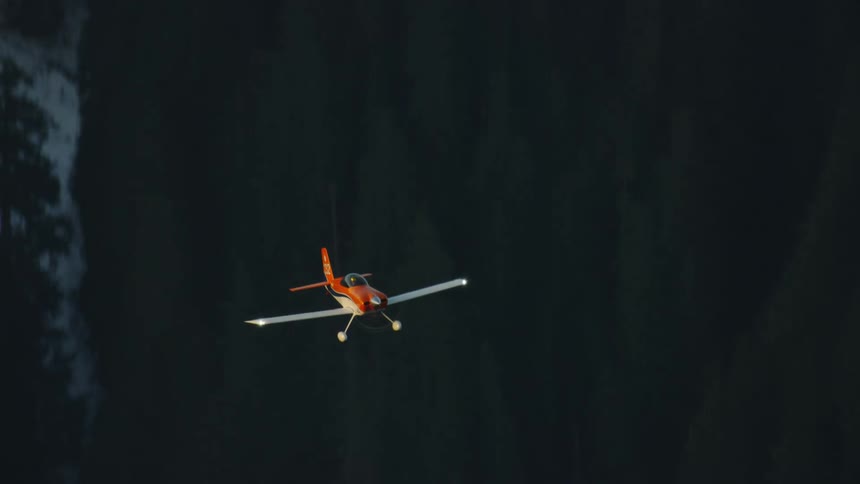 Air to air shot of an orange single engine engine acrobatic aircraft flying over the alpine forest