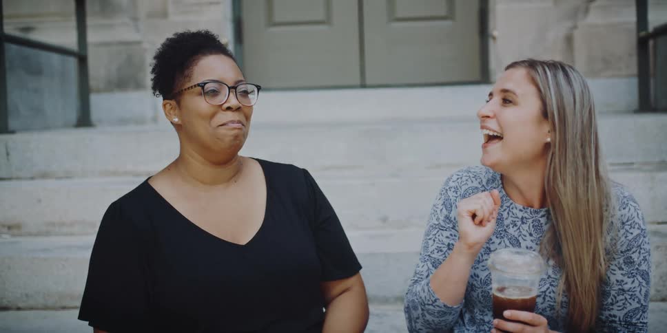 Two woman sit outside a building talking and holding drinks