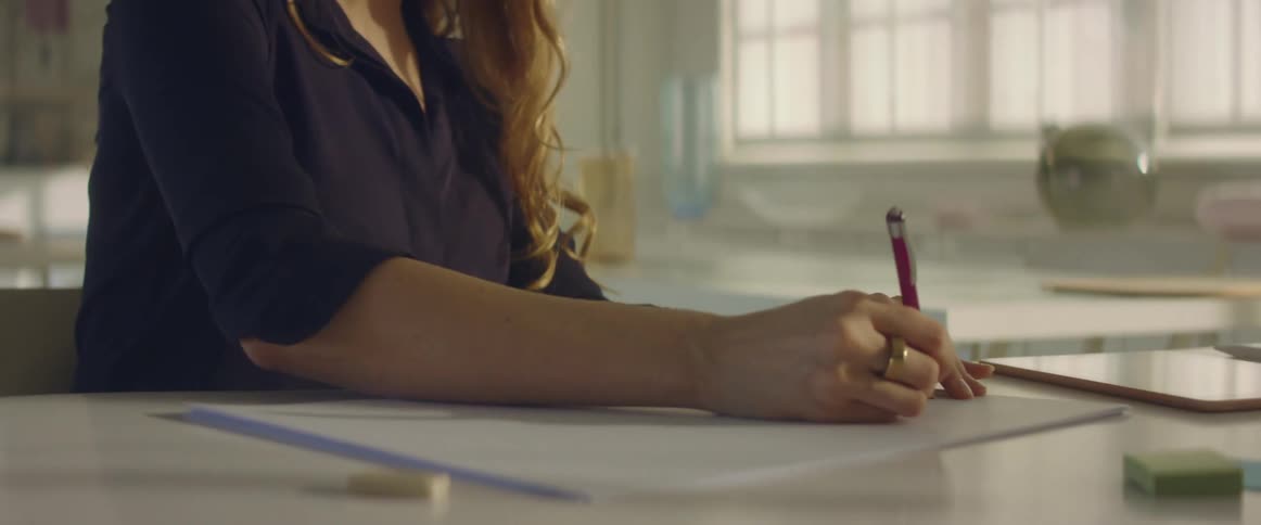 A woman sitting at a desk drawing on paper with a pen with a wall in the background