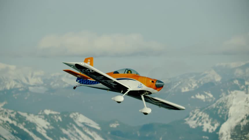 Close up shot of an orange and white single engine airplane rejoining the camera in formation flight high above the snow covered mountains in the winter