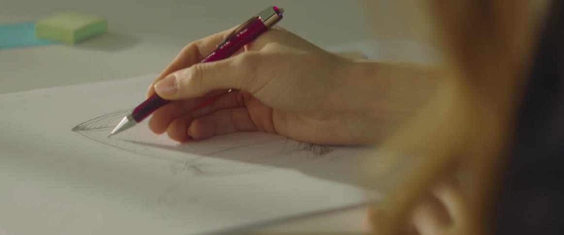 A woman in a room sitting at a desk drawing on a paper with a pen
