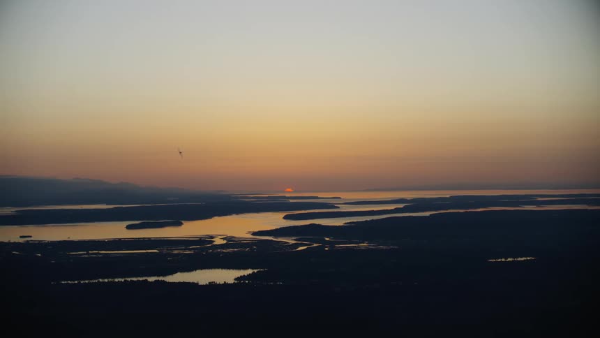 Super wide shot of an acrobatic airplane flying s turns in front of a setting sun and the Puget Sound Pacific Ocean as the sun sets