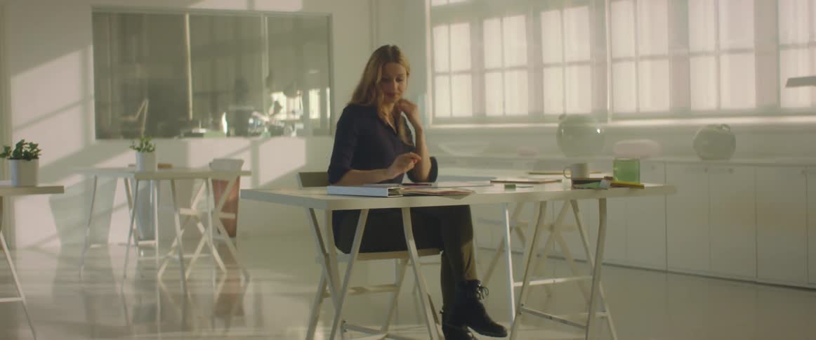 A woman sitting in a chair at a desk with a wall and windows in the background