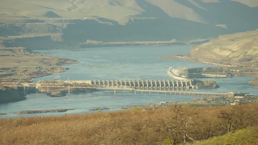 The Dalles Hydroelectric dam is seen through the branches of cherry trees in spring along the columbia river while an airplane lands in the background