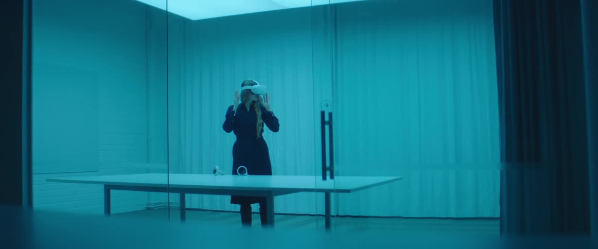 A woman lifts a virtual reality headset from a table and places it on her head while standing in a room