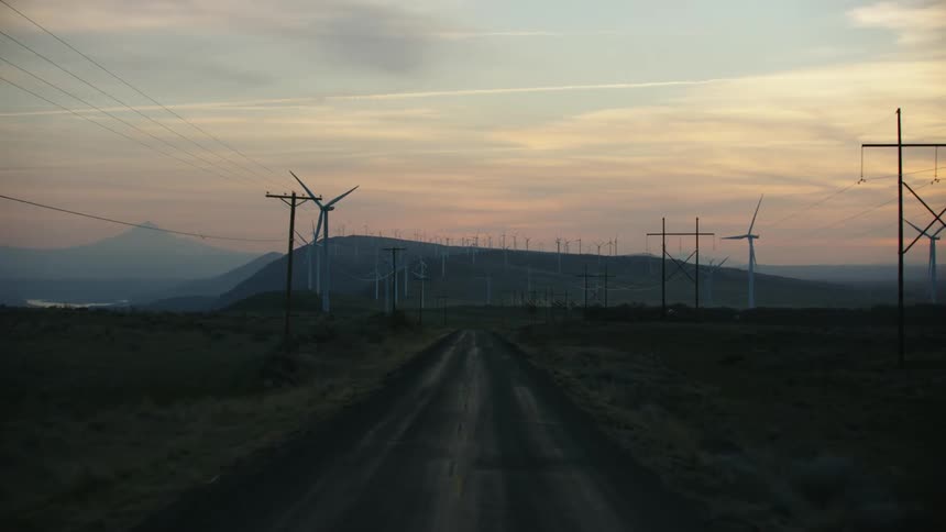 Wind turbines spinning along a rural road lined with turbines, power poles and a huge mountain can be seen on the distant horizon
