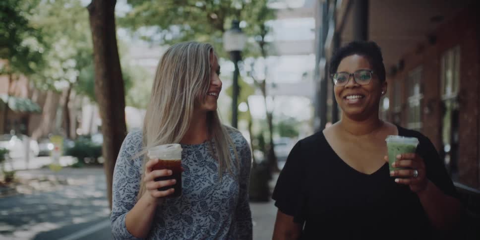 Two women walk down a town sidewalk together holding drinks