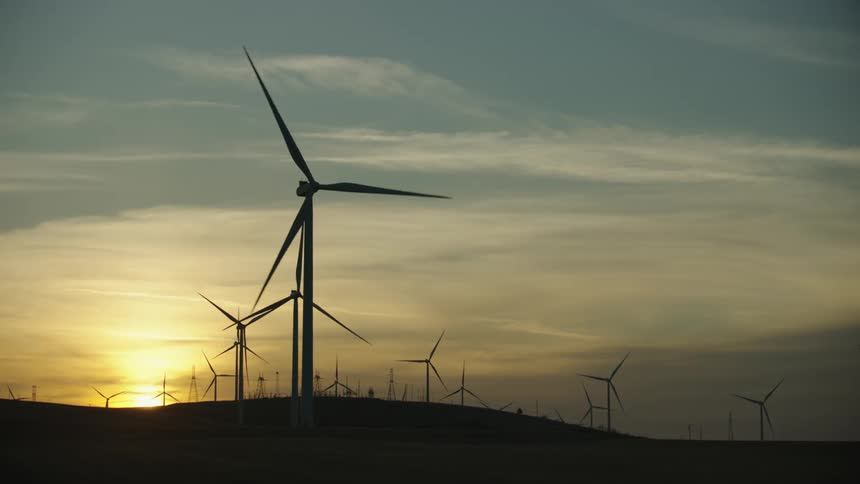 Wide statice shot of wind turbines spinning on a wind farm under a cloudy sky with the setting sun on the horizon