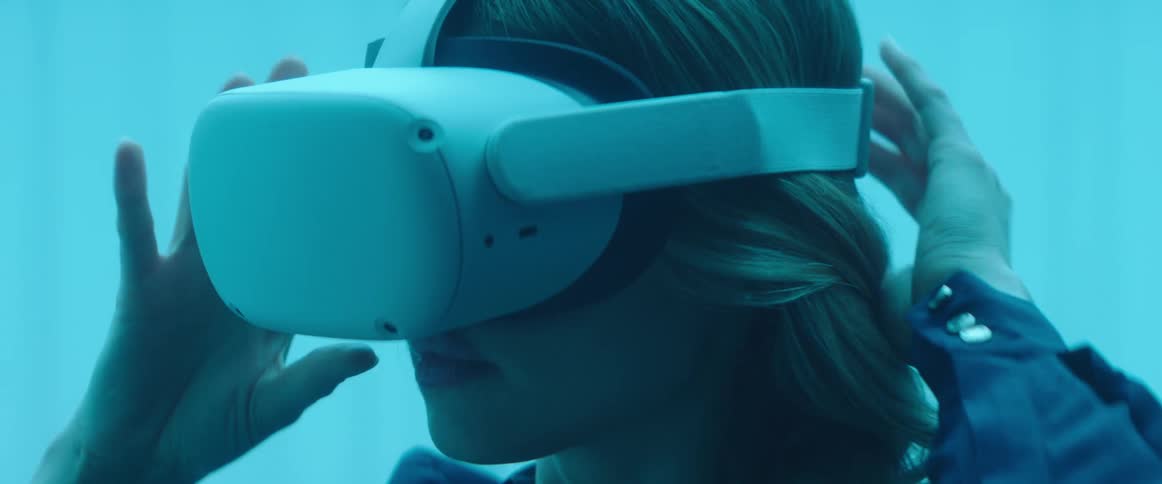 A woman in a room with a blue hue puts on a wide vr headset and then looks around