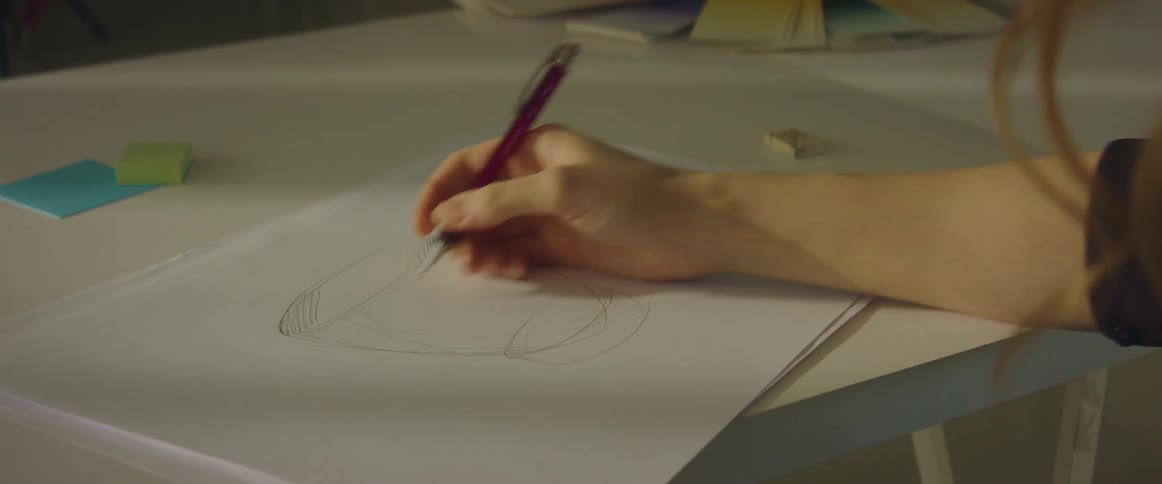 A woman in a room sitting at a desk drawing on a paper with a pen