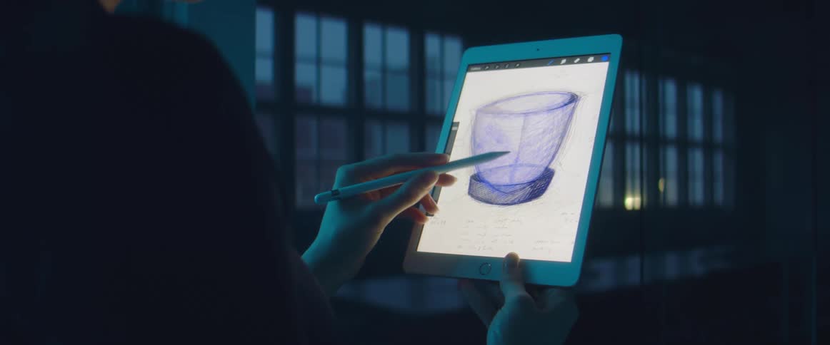 A woman holds a tablet computer an draws a sketch of a container on it using a stylus