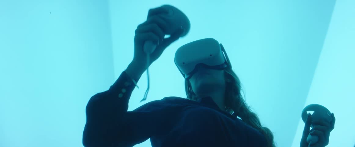 A woman wearing a virtual reality headset holds a controller which she waves around in front of herself