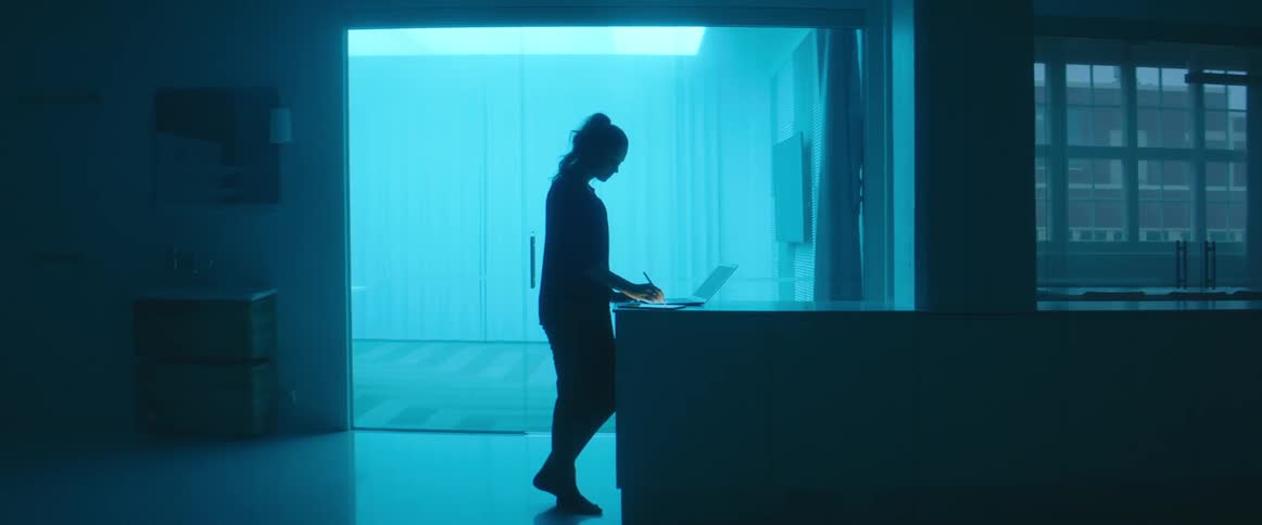The silhouette of a woman can be seen as she stands at a counter with a laptop on it