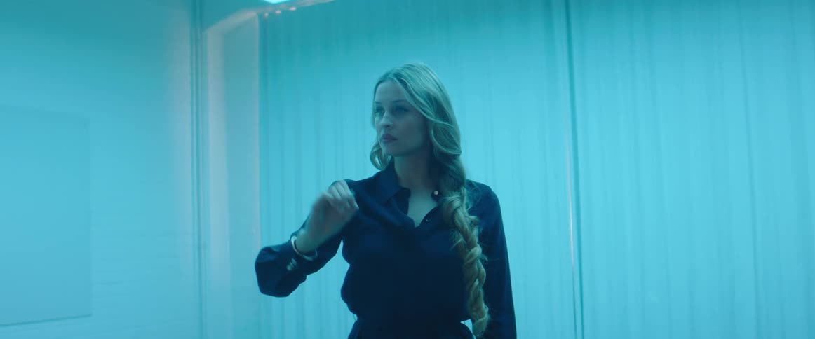 A woman stands in a blue lit room and pretends that she is writing on a wall