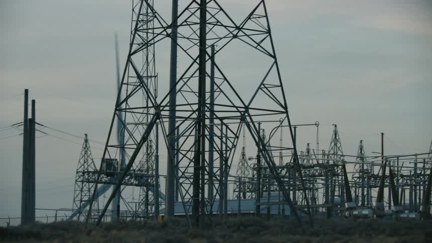 A power transfer substation with windmills turning in the background at sunset from the perspective of a passing vehicle