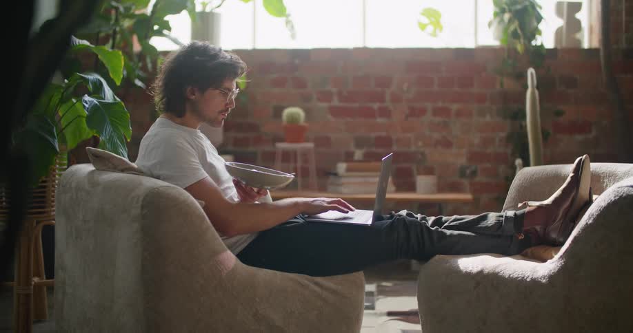 A man is typing on his keyboard in the living room with a bowl in his hand