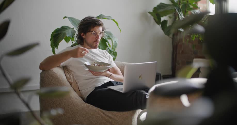 A man is eating food and looking at a laptop