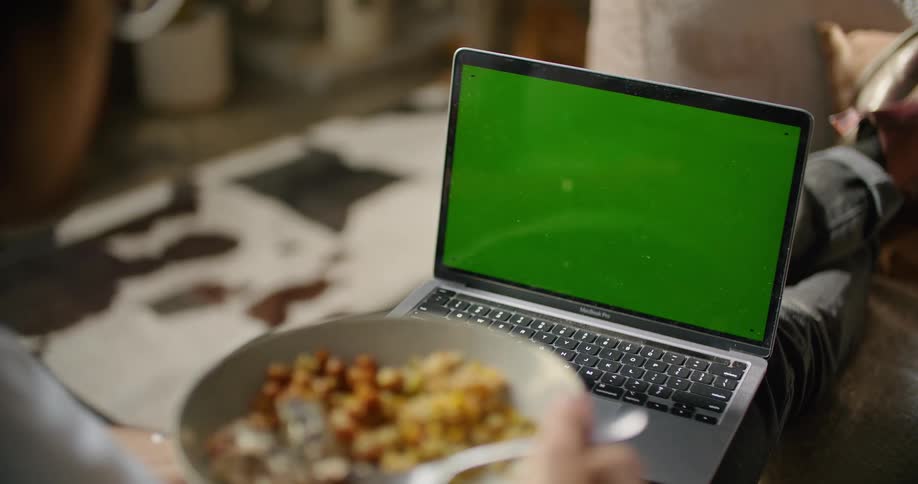 A person is sitting indoors and looking at a laptop while eating