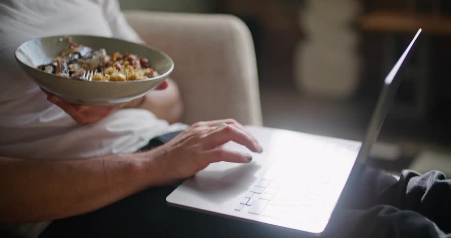 A man with glasses sitting on a couch holding a bowl of food and looking at a laptop