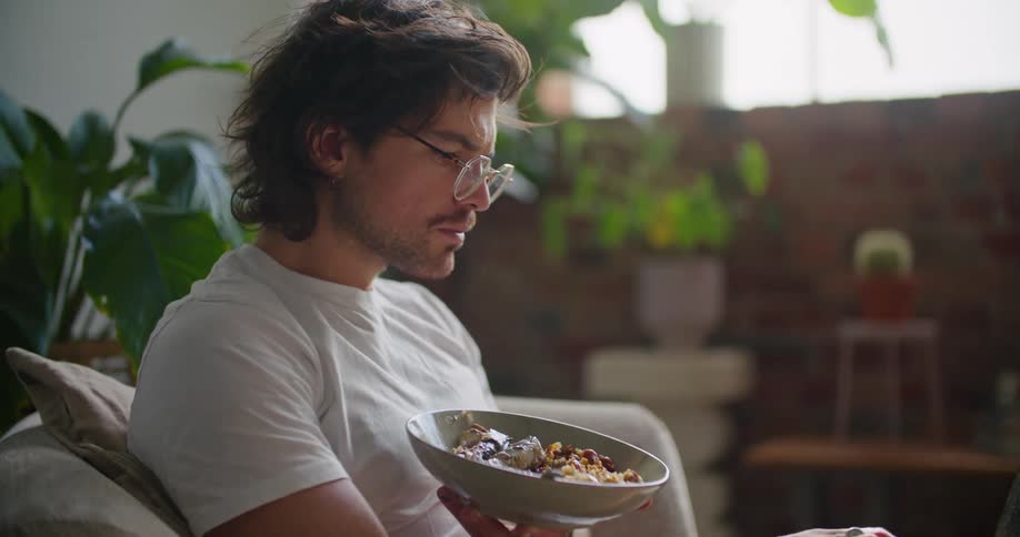 A man laying on a couch typing on a laptop and holding a bowl of food