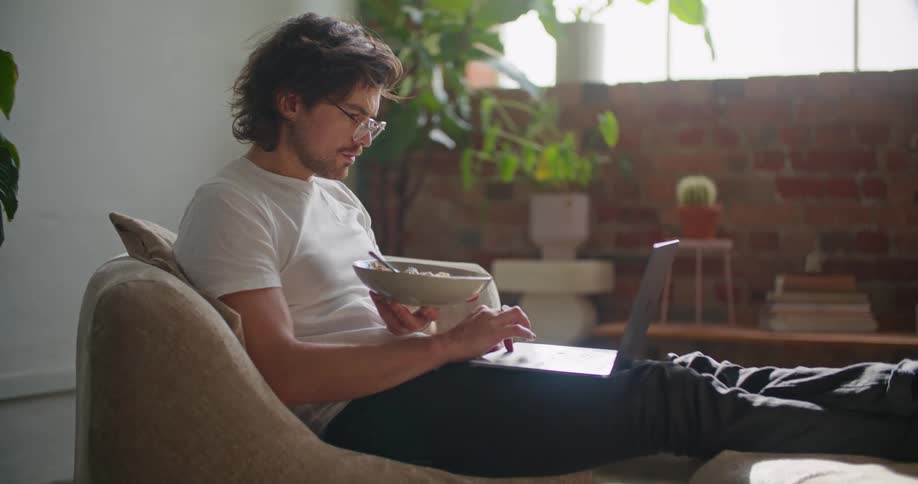 A man scrolls through his laptop while eating a bowl of cereal with his feet propped up on the couch