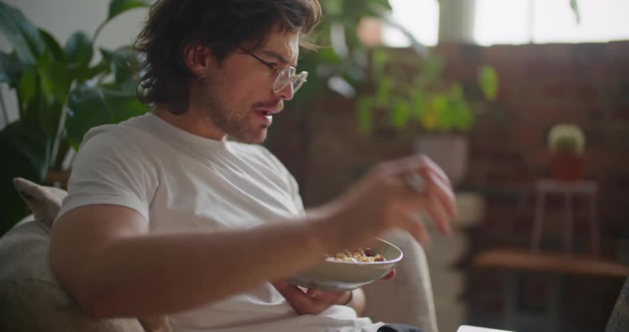 Man reclines on couch eating while closely examining contents of laptop screen