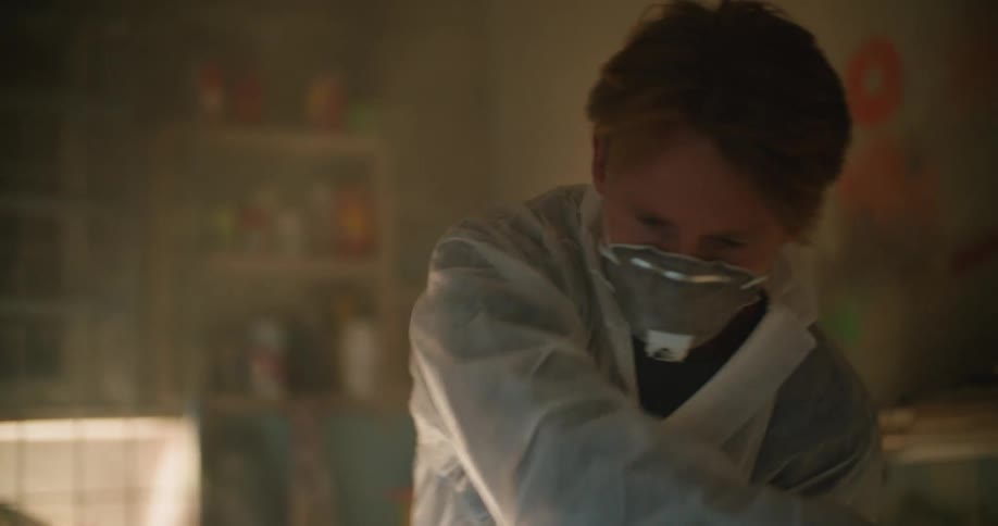 A man in a room with a mask sanding a surfboard with a wall and window in the background
