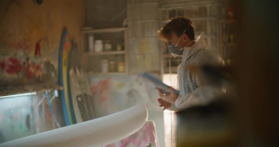 A man in a face mask and protective suit checks on a surfboard hes building