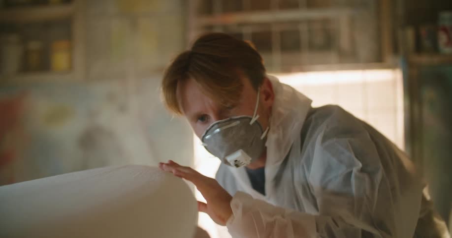 A man in a room with a mask sanding a surfboard with a wall and window in the background