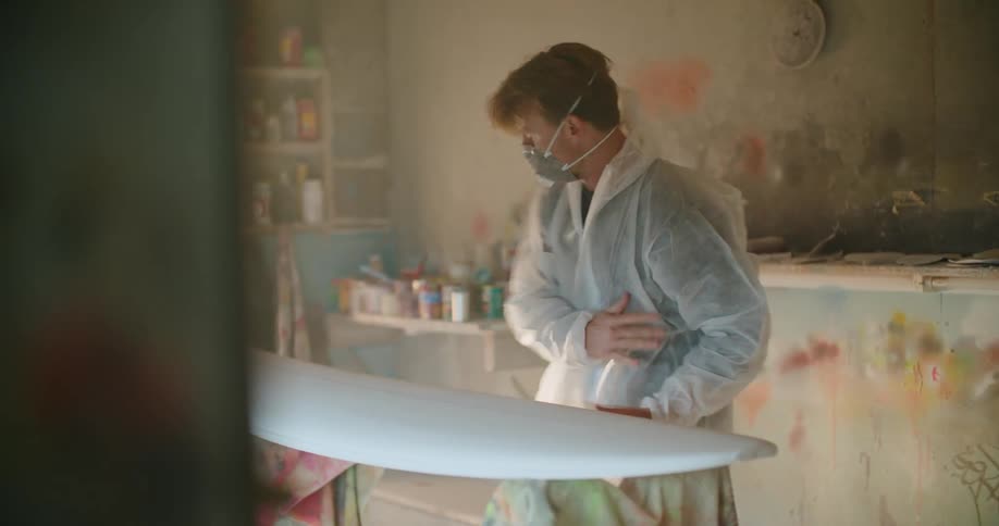 A man wearing a mask and coveralls carries a board into a workshop to sand and paint