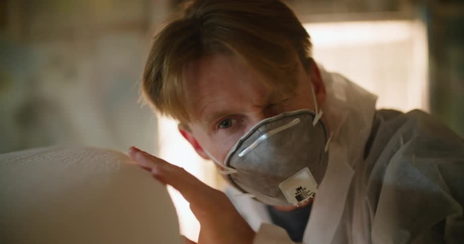 A man wears a face mask and protective suit as he sands a large piece of wood