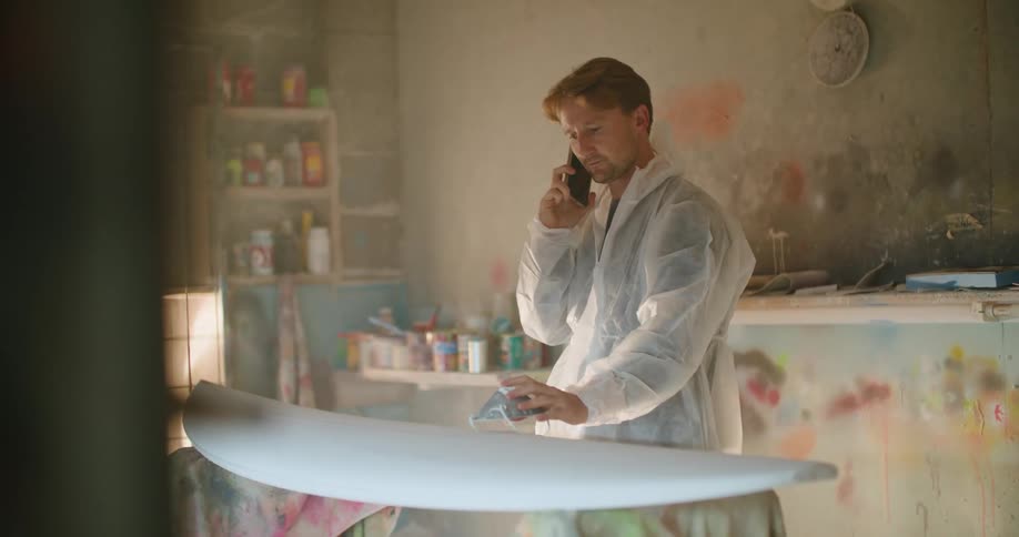 A man is talking on the cell phone as he works on a surfboard