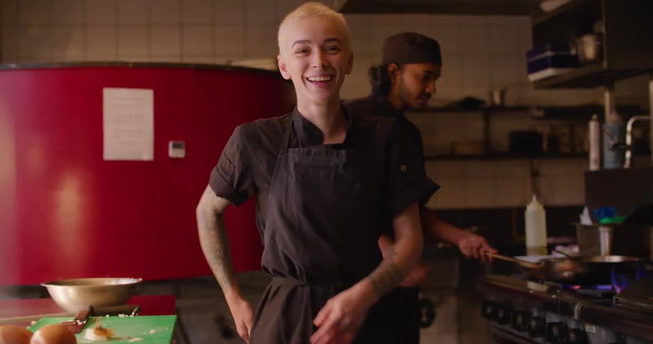 A kitchen chef laughs as she watches a colleague cook over a stovetop