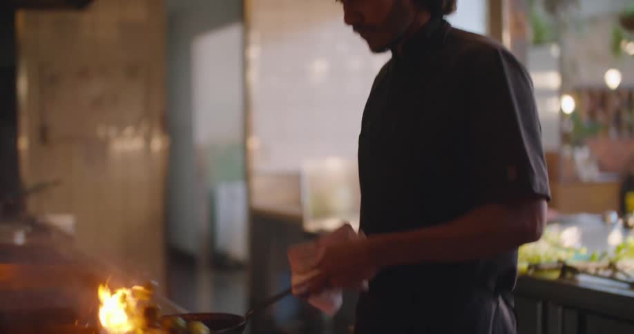 A man with a hat cooking food in a frying pan over a stove with fire and smoke