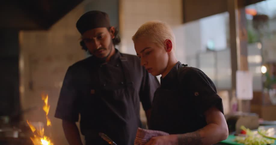 A restaurant chef cooks over a large fire in the kitchen beside another chef watching