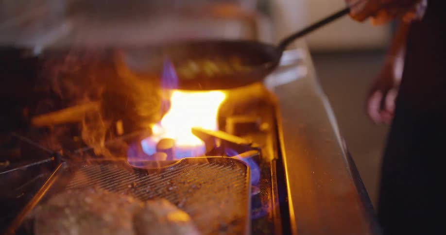 A man flips a pan with vegetables in it as meat grills