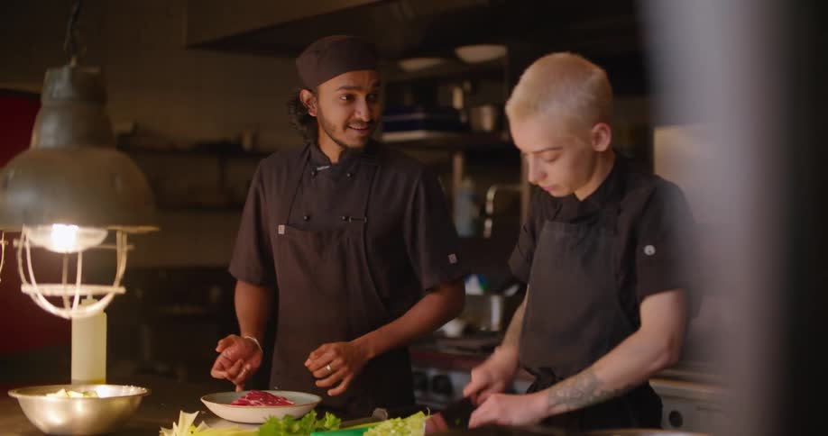 Two restaurant chefs talk together in the kitchen while one of them is chopping food