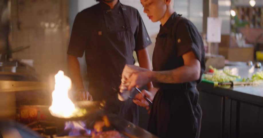 Two chefs in a restaurant kitchen cooking food over a flame in a pan on a stovetop