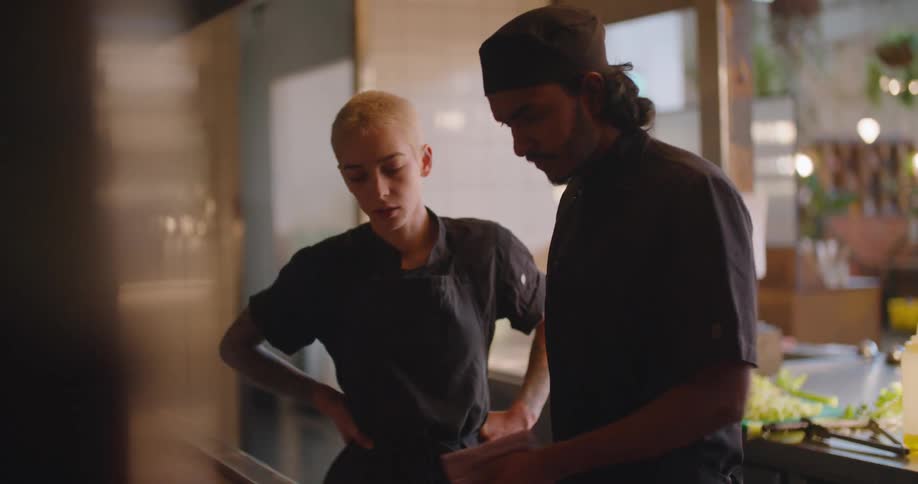 A chef flips food in a pan over an open fire while another chef watches him