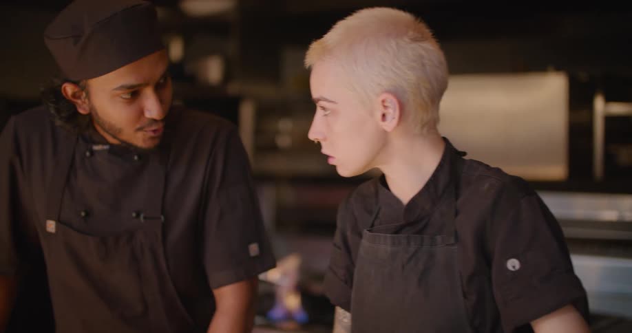 A man and a woman with aprons in a kitchen talking with fire on a stove in the background