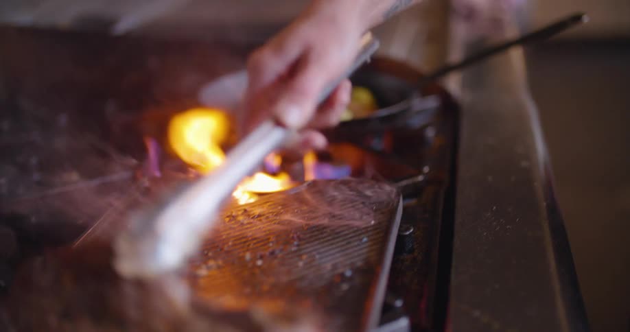 A person in a kitchen grilling steak with fire and smoke