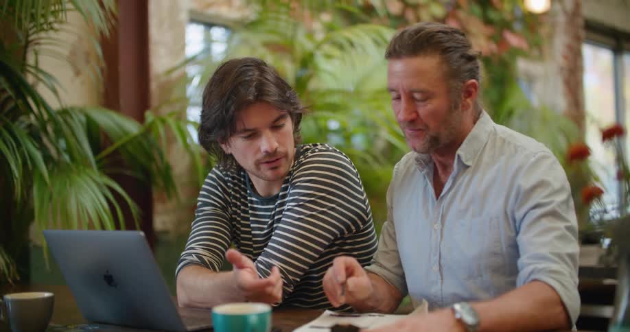 Two men sit together at an outside table looking at a paper and talking about it